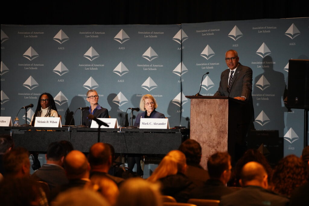 Executive committee sits behind a table on a stage while Mark Alexander stands at a podium in front of an AALS backdrop