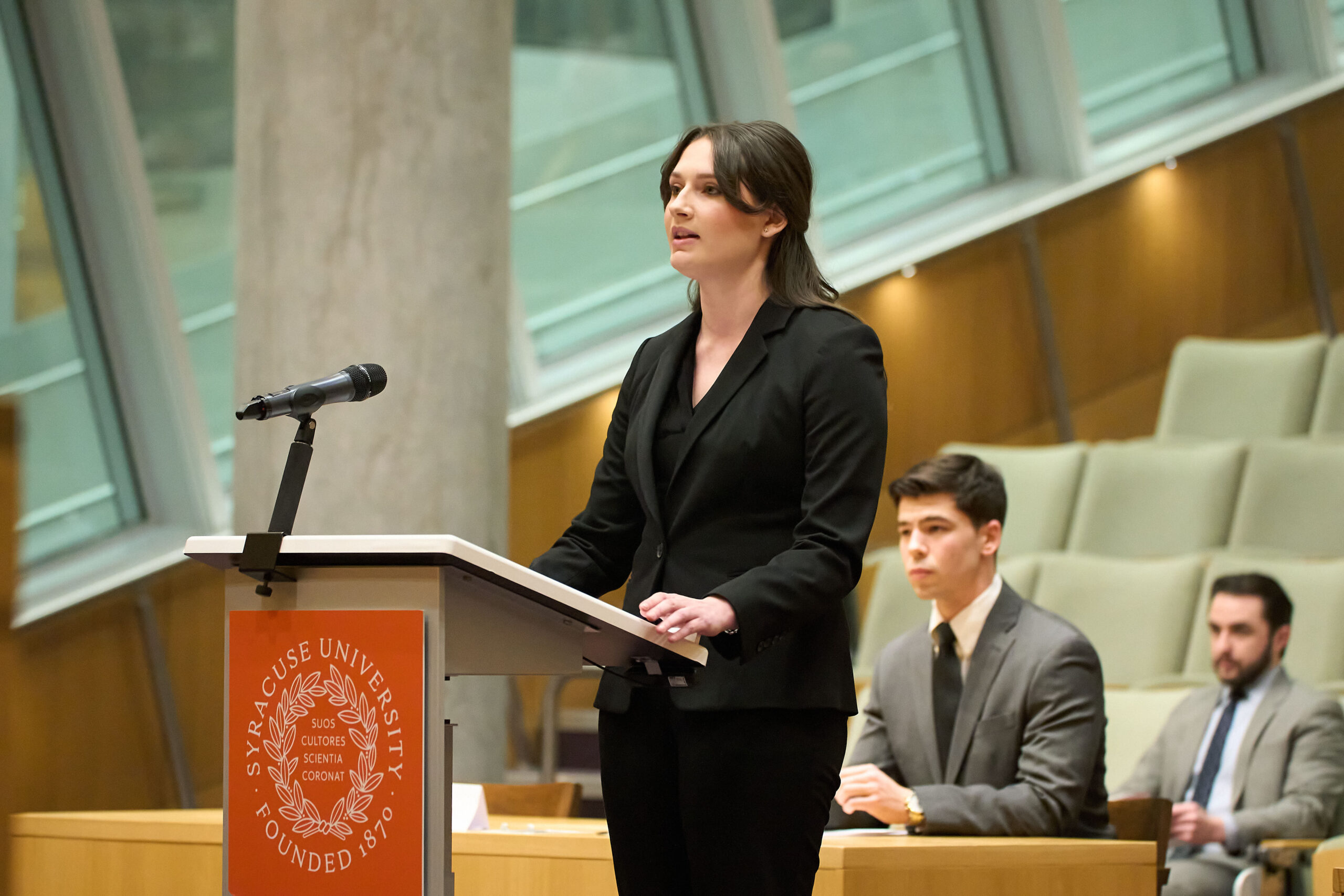 Syracuse University College of Law students participating in its Advocacy Program with student standing in a black suit at podium speaking, another student sitting at a desk and a person in the audience listening.
